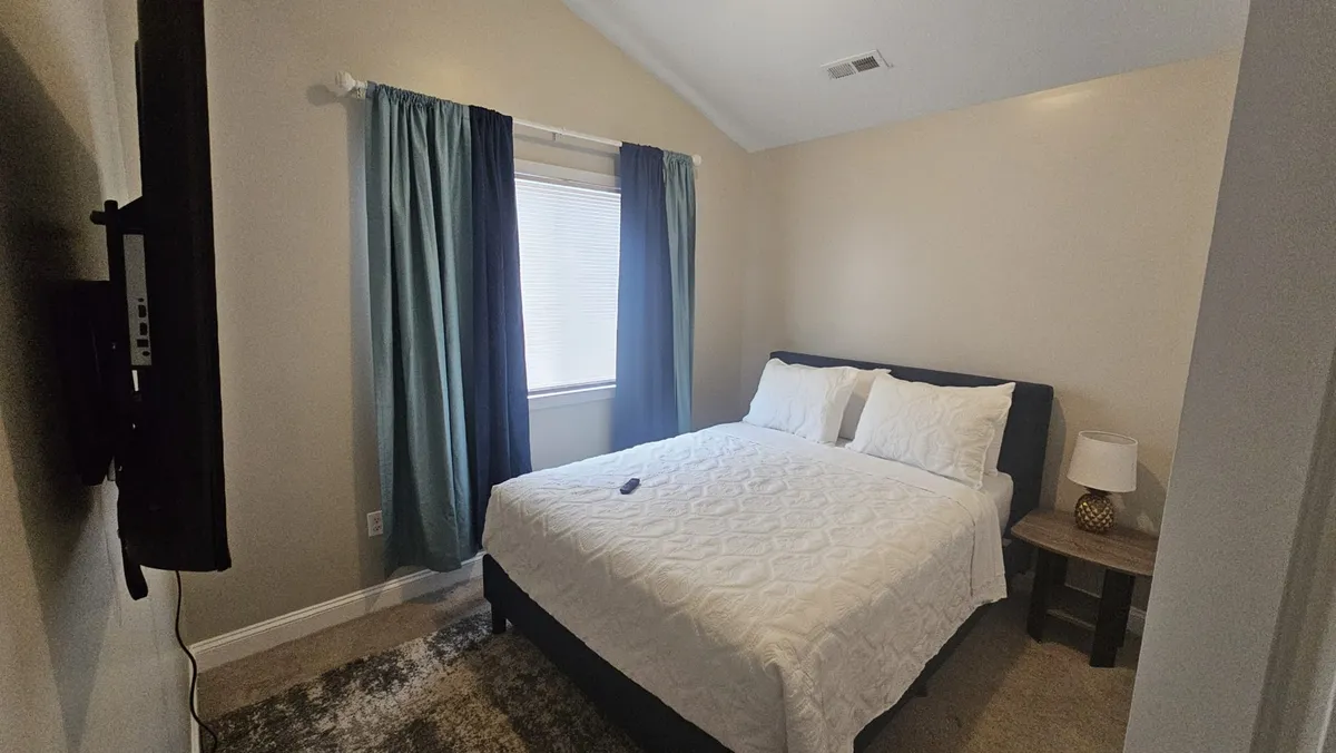 A warmly lit bedroom with a neatly made bed positioned next to a large window, soft curtains, a nightstand, and a glowing lamp. The space invites relaxation and a peaceful atmosphere, ideal for unwinding after a day of activities.