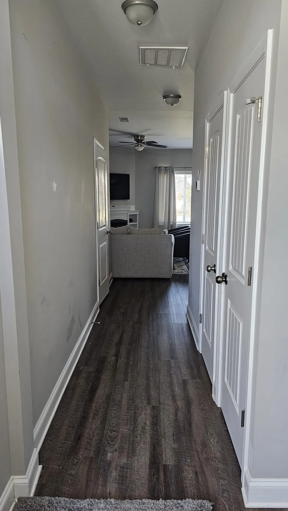 This image captures a long hallway leading directly into a bright, inviting living room. The living area is visible at the end, suggesting an open layout that welcomes guests and encourages togetherness as soon as you enter the home. A decorative ceiling fixture adds a touch of style, while the view hints at shared moments and gatherings.