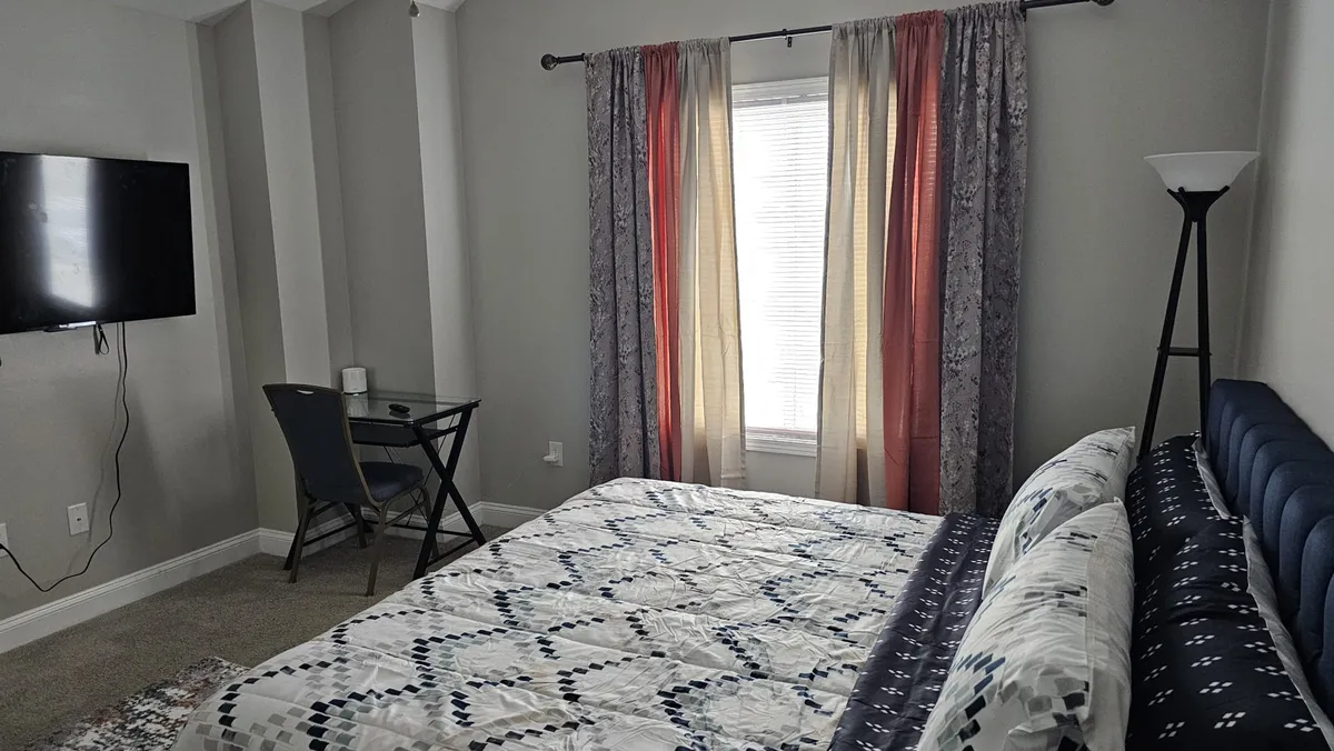 A calm and inviting bedroom featuring a comfortable bed, a window with a view, a functional desk, and a smart TV—offering versatile space for both relaxation and group movie nights.