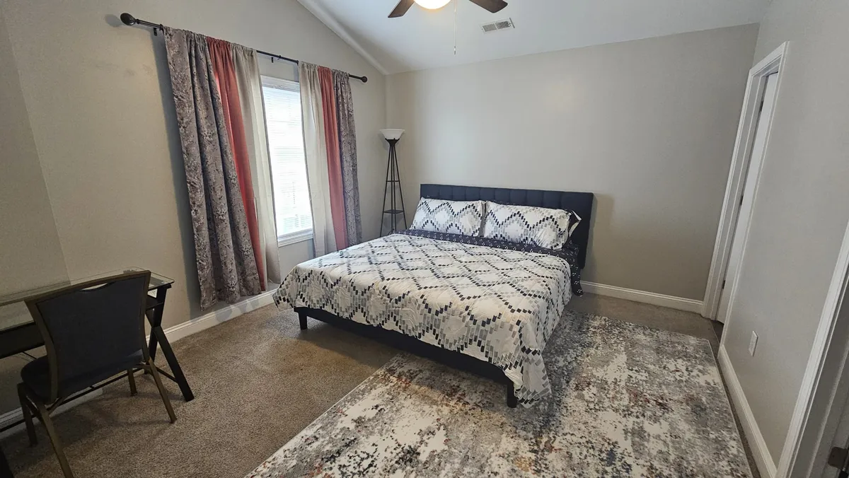 A serene bedroom scene featuring a comfortable bed, elegant curtains, a soft rug, gentle lighting from a bedside lamp, and a functional desk by the window. The space shows versatility for both relaxation and productivity, offering options to work, read, or simply relax.