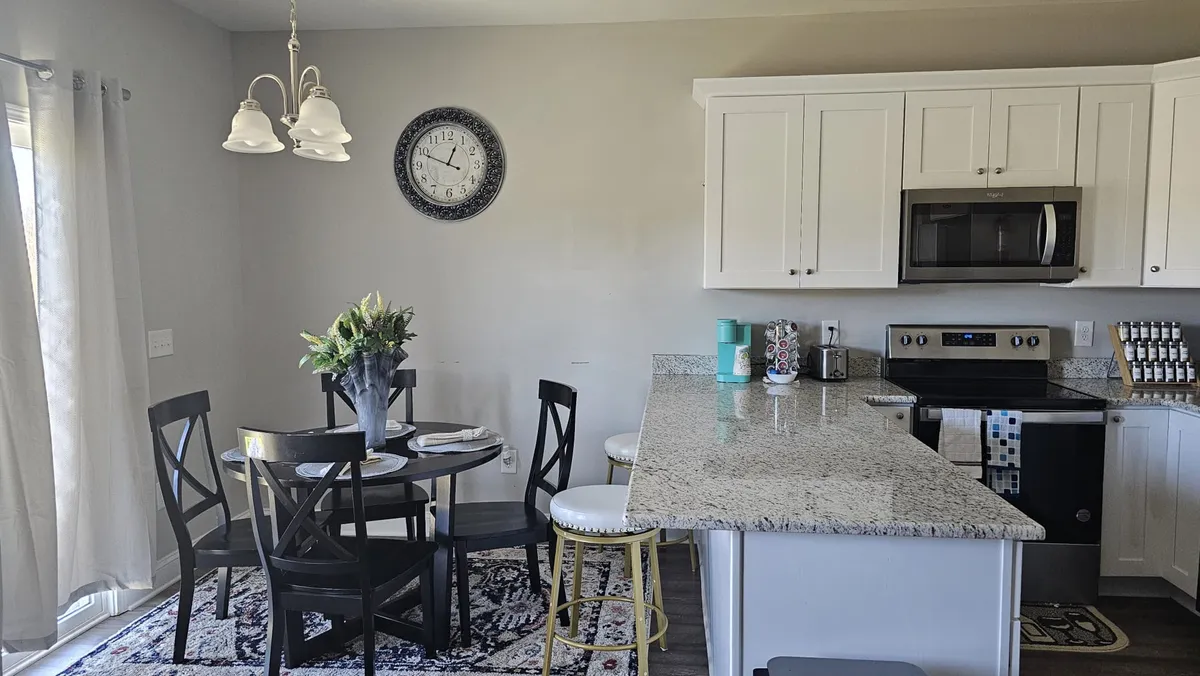 A welcoming dining area adjacent to the kitchen, highlighted by a sturdy dining table, a kitchen island, a wall clock, and a floral centerpiece. The atmosphere is warm and inviting, set up for shared meals, conversations, and connecting with friends and family.