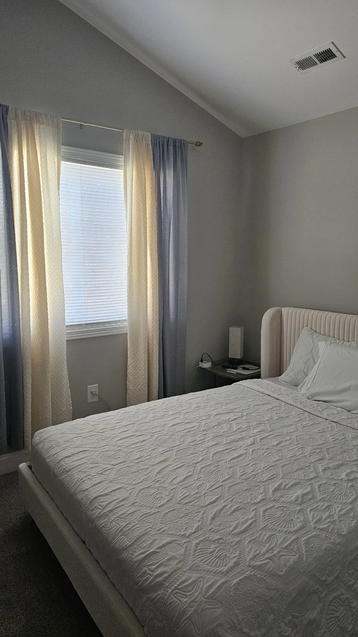 A tranquil bedroom filled with natural light, featuring a comfortable bed, nightstand, and curtains framing a window. The peaceful atmosphere suggests a restful environment for families.