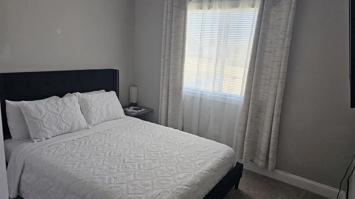 This image features a bedroom with a plush bed positioned near a window, complemented by light curtains that let in natural light. The bed is made with quality linens, and the tranquil setting suggests a space perfect for resting, reading, or unwinding after a long day.