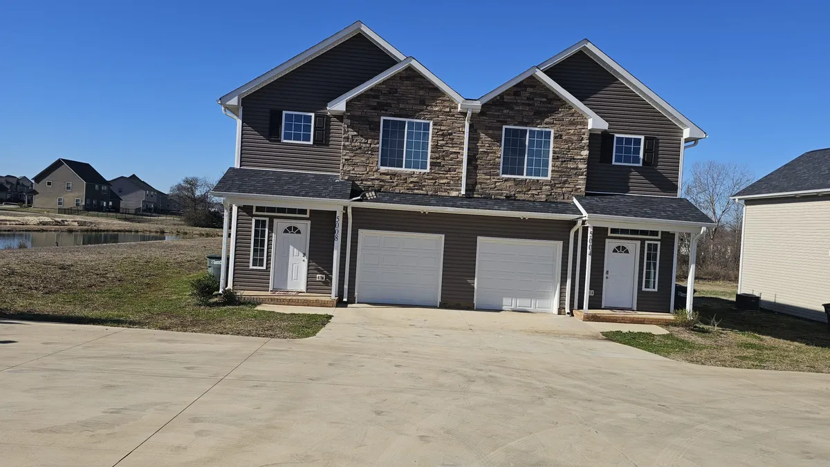 The image showcases the inviting exterior of a modern duplex in Concord, North Carolina, featuring elegant stone accents, two front doors, and a spacious driveway perfect for family arrivals. The well-maintained facade and open front yard suggest an ideal spot for outdoor gatherings, barbecues, and children playing, with a welcoming atmosphere that feels perfect for milestone celebrations and building memories.